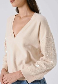 Maglione beige chiaro con scollo a V e maniche lunghe. Le maniche presentano un tessuto strutturato con paillettes. Polsini a costine e vestibilità rilassata.