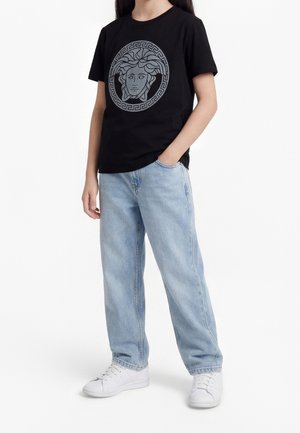 Osoba nosząca czarną koszulkę z logo Medusa, jasnoniebieskie dżinsy i białe tenisówki, stojąca z jedną ręką w kieszeni.