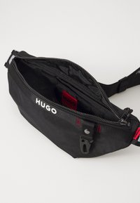 Juodas audinys liemuo krepšys su užtrauktuku, su "HUGO" logotipu, raudonais akcentais ir reguliuojama juosta nešimui.