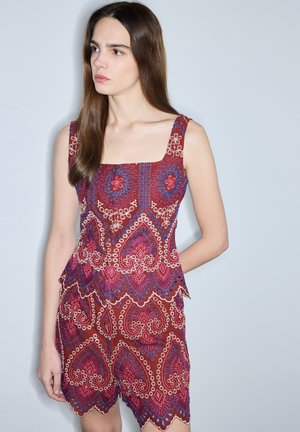 Junge Frau mit langen braunen Haaren, die ein ärmelloses rotes Kleid mit Muster und wellenförmigen Rändern trägt, schaut nach links vor einem schlichten hellen Hintergrund.