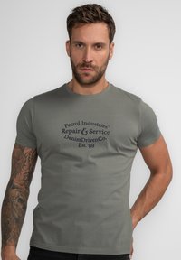 Oliven grøn bomulds t-shirt med korte ærmer og rund hals. Har et mørkeblåt trykt logo på forsiden. Glat overflade.