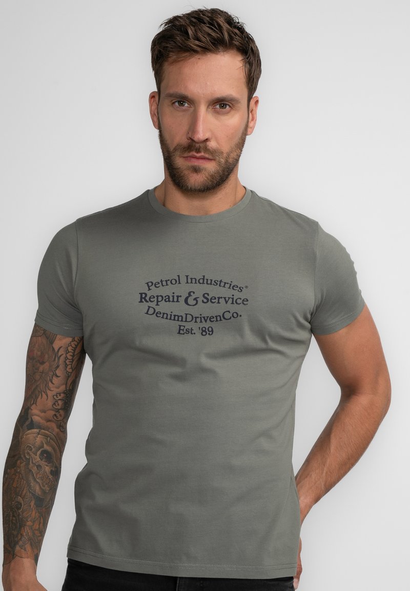 Oliven grøn bomulds t-shirt med korte ærmer og rund hals. Har et mørkeblåt trykt logo på forsiden. Glat overflade.