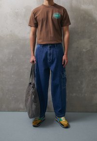 Hnědé bavlněné tričko s tyrkysovým logem, modré denimové cargo kalhoty, šedá látková taška a žluté tenisky s modrými akcenty. Viditelné texturované povrchy.