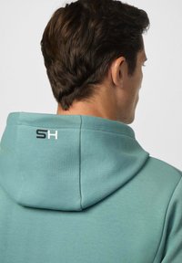 Sudadera azul claro con una textura suave y capucha, que presenta un pequeño logo 'SH' en la parte posterior. Tejido suave y corte relajado.