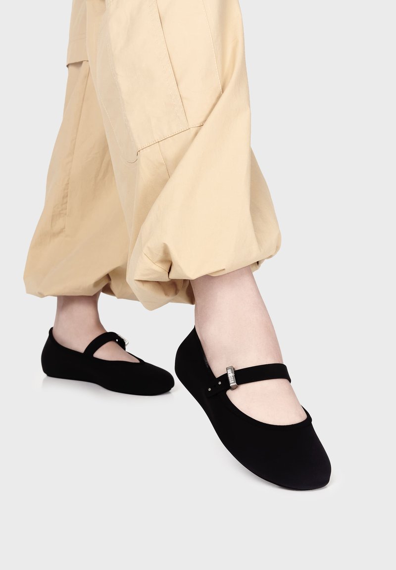 Bailarinas negras con una textura suave y mate y una correa en el pie, realzada por una hebilla plateada. Llevadas con pantalones beige claros y recogidos.