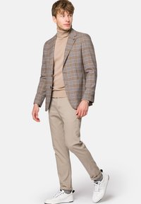Brązowa kratkowana marynarka na beżowym golfie, w połączeniu z jasnobeżowymi spodniami i białymi sneakersami. Marynarka ma jeden guzik i klapy.