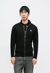 Juodas megztinis su užtrauktuku, pagamintas iš megzto audinio, su raištuotomis rankovėmis ir maža logotipo žyme krūtinės srityje. Dėvimas ant marginto marškinėlio.