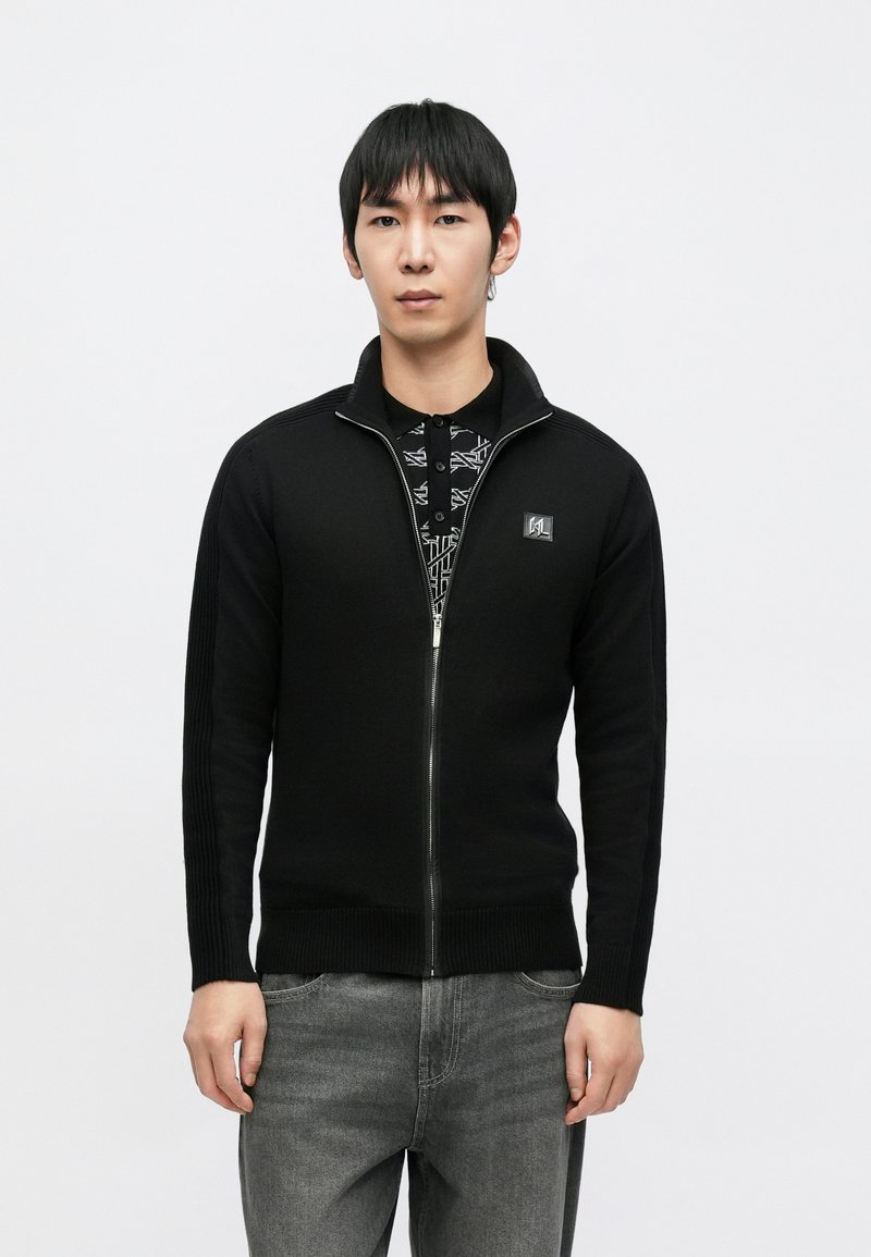 Juodas megztinis su užtrauktuku, pagamintas iš megzto audinio, su raištuotomis rankovėmis ir maža logotipo žyme krūtinės srityje. Dėvimas ant marginto marškinėlio.