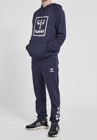 Person mit navyblauem Hummel-Hoodie und passender Jogginghose, trägt Sneaker und steht mit den Händen in der Hoodie-Tasche vor einem einfarbigen Hintergrund.