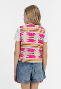 Multifarebný tkaný nátelník s ružovými, oranžovými a modrými vzormi, s textúrovaným povrchom. Nosený cez bielu košeľu a v kombinácii s džínsovými šortkami.