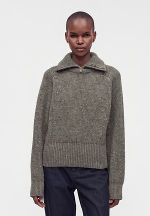 Persona que lleva un suéter gris de punto con cuello con cremallera y vaqueros azul oscuro, de pie frente a un fondo liso y claro.