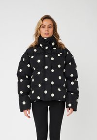 Svart pufferjakke med oversized passform, med store hvite prikkete m�ønstre, høy krage og elastiske mansjetter. Teksturen ser glatt og blank ut.
