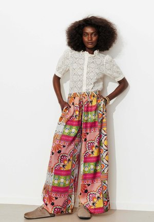 Femme aux cheveux bouclés et volumineux porte un chemisier blanc en dentelle à manches courtes et un pantalon à jambes larges avec des motifs floraux et géométriques éclatants.