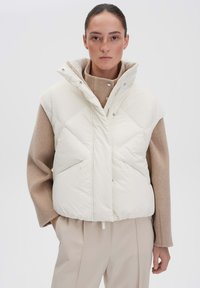 Weißer Taillengilet mit hohem Kragen und ärmellosem Design, mit Rautensteppung und zwei Fronttaschen, kombiniert mit einem beigen Pullover.