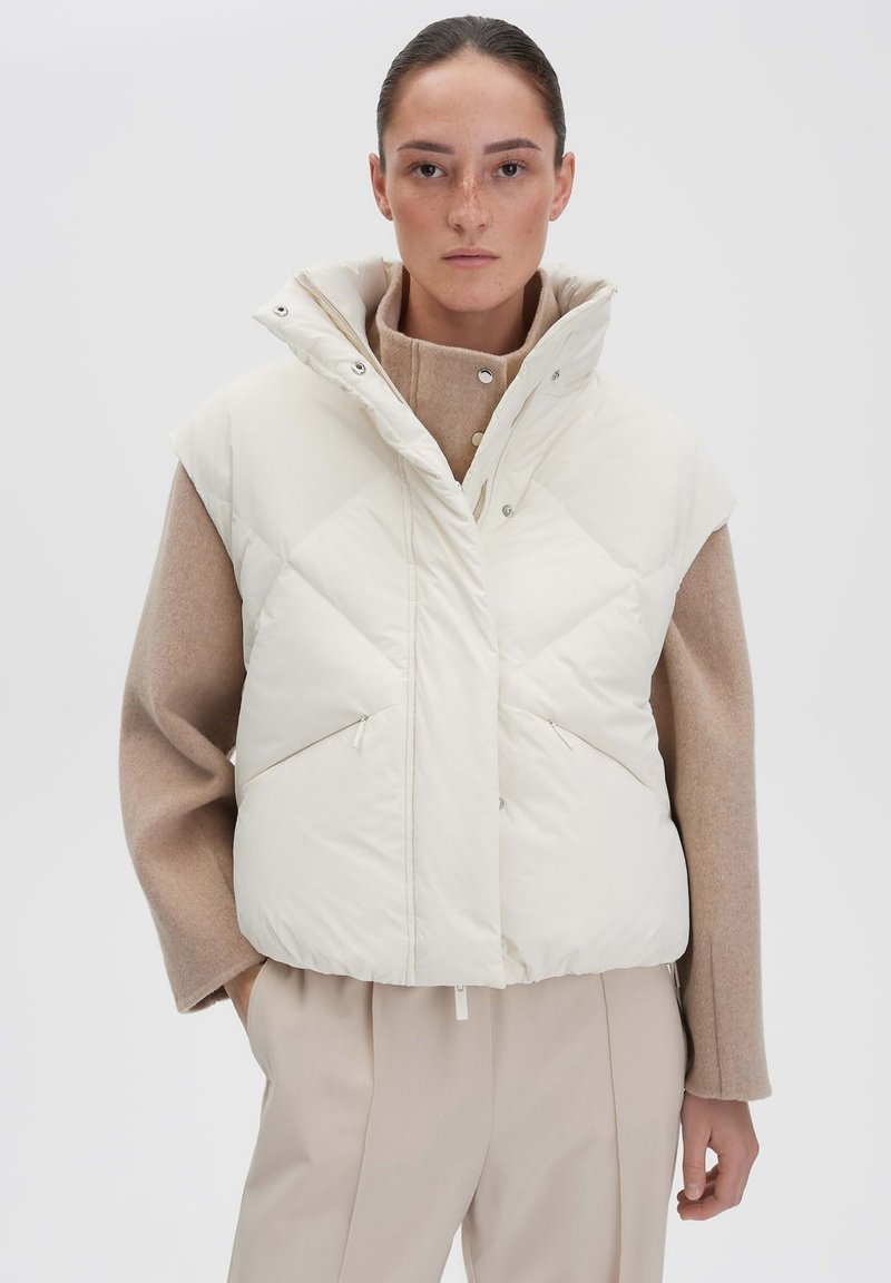 Weißer Taillengilet mit hohem Kragen und ärmellosem Design, mit Rautensteppung und zwei Fronttaschen, kombiniert mit einem beigen Pullover.