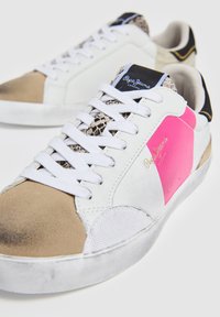Zapatilla blanca con acentos en rosa y beige, panel de textura de serpiente, cordones blancos y suela de goma; logo en la lengüeta.