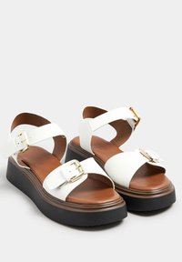 Sandali wedge bianchi in pelle con piattaforma marrone. Presentano cinturini incrociati, fibbie regolabili e una superficie liscia. Accenti in hardware dorato.