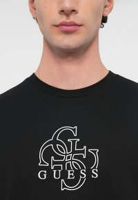 Melna kokvilnas T-kreklu ar baltu logotipa dizainu, kurā ir sapītu burti un teksts "GUESS". Īsas piedurknes un apaļa apkakle.