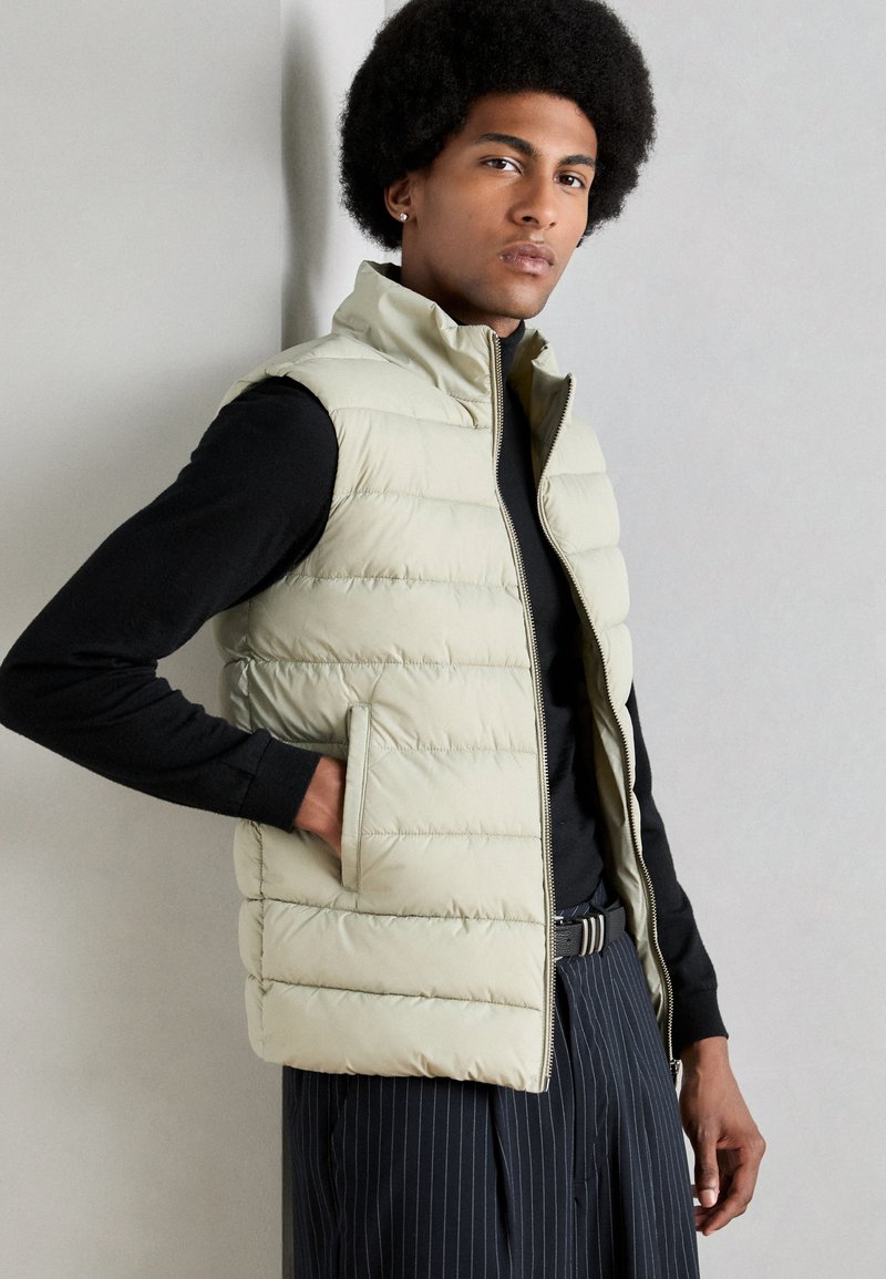 Young man with afro hairstyle wearing beige quilted vest over black long sleeve shirt and pinstriped trousers, standing against plain background.
