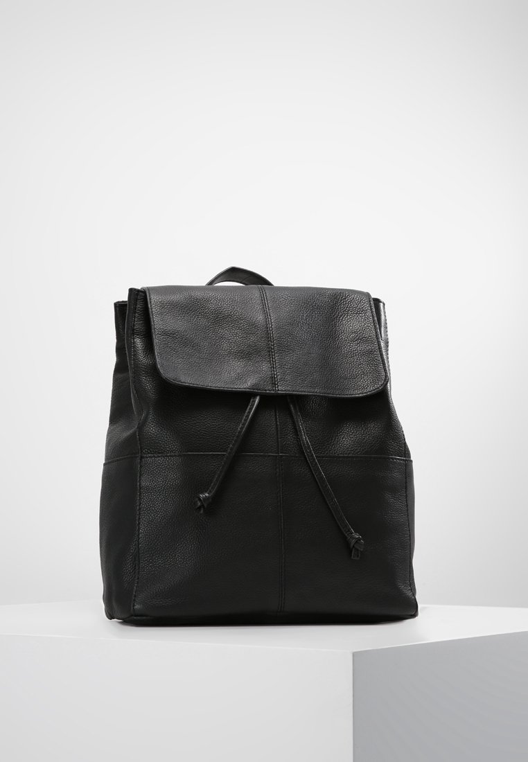 Sac à dos en cuir noir avec fermeture à rabat et cordon, présenté sur un socle blanc devant un fond gris uni.
