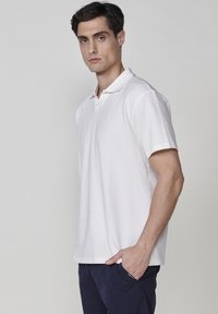 Vit polo-shirt med texturerad tyg, korta ärmar och platt krage, med en subtil logotypdetalj på bröstet.