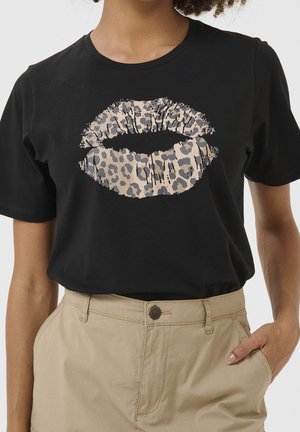 Czarny T-shirt z dużym nadrukiem w kształcie ust w panterkę, noszony z beżowymi spodniami z wysokim stanem, ręka w kieszeni.