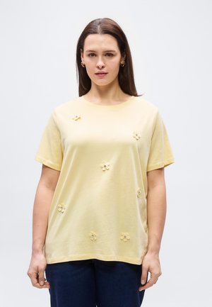 Femme aux cheveux bruns raides portant une chemise jaune pâle à manches courtes ornée de petites fleurs en tissu, associée à un pantalon foncé, sur fond blanc.