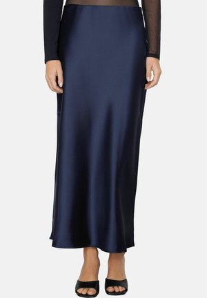 Frau trägt einen marineblauen Satin-Midirock kombiniert mit schwarzen offenen High Heels und einem durchsichtigen schwarzen Langarmoberteil.