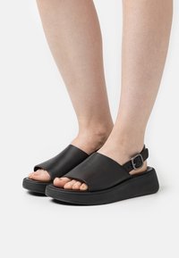 Musta avonainen sandaalit, jotka on valmistettu sileästä tekonahasta. Niissä on leveä hihna ja säädettävä nilkkakiinnitys, ja ne ovat vaahtomuovipohjalla.