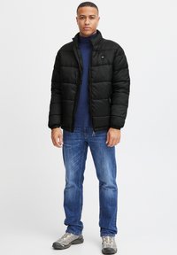 Svart pufferjakke med ståkrage og quilting design, båret over en blå genser med høy hals, kombinert med blå jeans og grå sneakers.