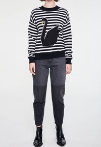 Czarnobiały sweter w paski z motywem czarnej łabędzicy, noszony z ciemnoszarymi, zwężanymi dżinsami i botkami za kostkę. Sweter ma ściągacze przy mankietach.