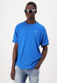 Μπλε βαμβακερό T-shirt με γυρισμένα μανίκια και μικρό κίτρινο λογότυπο. Συνδυασμένο με ανοιχτό μπλε τζιν. Μοντέλο που φορά λευκά γυαλιά ηλίου.