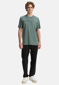 Grøn t-shirt med korte ærmer og rund hals, med en sort vandret stripe hen over brystet, parret med sorte bukser og hvide sneakers.