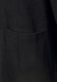 Gros plan d’un tissu côtelé noir avec une poche horizontale cousue en surface, montrant la texture tricotée et l’ouverture de la poche.