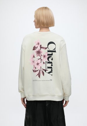 Osoba z krótkimi blond włosami nosi białą bluzę z różowym wzorem kwiatów wiśni i dużym napisem "Cherry Blossom" na plecach.