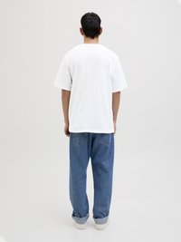 Weißes T-Shirt mit kurzen Ärmeln, lockere, blaujeans mit hochgekrempelten Säumen und weiße Sneaker, von hinten gesehen vor einem neutralen Hintergrund.