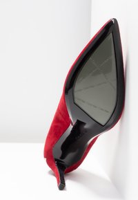 Escarpin en daim rouge à talon haut avec un bout pointu et une semelle en cuir laqué noir, présentant le nom de la marque embossé sur la semelle.