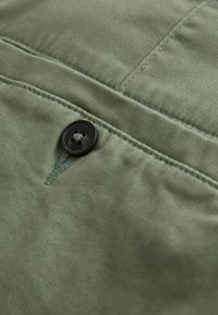 Gros plan d'un tissu vert olive avec des coutures détaillées et un bouton marron foncé à quatre trous cousu près d'une boutonnière.