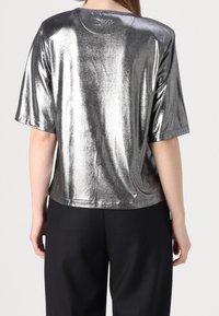 Top corto a maniche corte di colore argento metallico con una vestibilità ampia e scollo rotondo. Presenta una texture riflettente e un logo discreto sul retro.