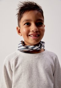 Jeune garçon souriant aux cheveux en désordre, portant un sweat-shirt gris clair et une écharpe rayée noir et blanc autour du cou, sur un fond uni.