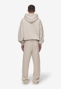 Hoodie oversized bege com capuz forrado, ombros caídos e punhos canelados, combinado com calças de treino largas a condizer. Tecido suave e texturizado.