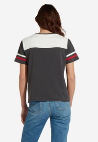 Šedé tričko s krátkým rukávem a bílým horním panelem, s červenými a bílými pruhovanými akcenty na rukávech. Měkký bavlněný materiál.