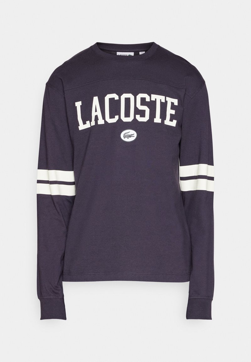 Marineblauw overhemd met lange mouwen met "LACOSTE" in witte letters, voorzien van een logo-patch en witte strepen op de mouwen. Zachte stof, ontspannen pasvorm.