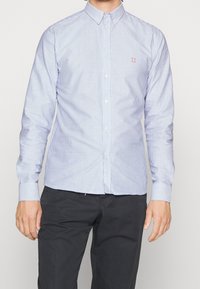 Camicia button-up a righe azzurre e bianche con colletto, dotata di maniche lunghe e un piccolo logo rosso sul petto. Tessuto dalla texture liscia.