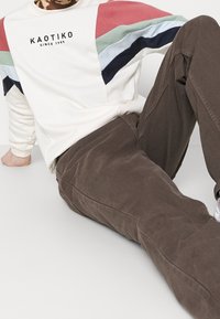 Valkoinen collegepaita, jossa on punaiset, vihreät ja tummansiniset yksityiskohdat ja painettu logo. Yhdistetty ruskeisiin, kuvioituihin housuihin.