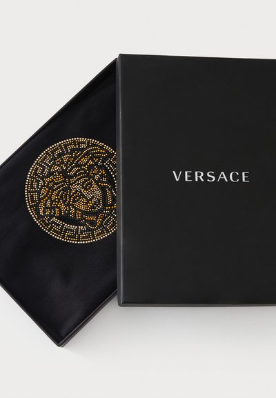 Juoda Versace dovanų dėžutė, kurioje matomas išraiškingas aukso ir sidabro detaliai puoštas raštas su apvaliu raštu ant juodos audinio vidinės dalies.