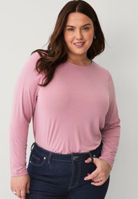 Long-sleeve top in soft pink fabric with a round neckline. Paired with dark blue denim jeans featuring a button and zip closure.
