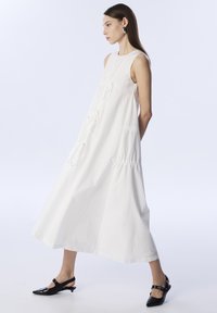 Vestido sin mangas de algodón blanco con detalles fruncidos, una silueta fluida y lazos laterales; combinado con tacones de salón negros.
