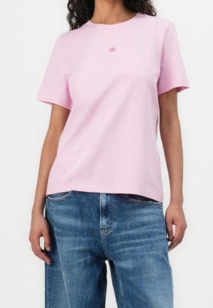 Femme portant un t-shirt rose clair à manches courtes avec un petit logo sur la poitrine et un jean bleu.