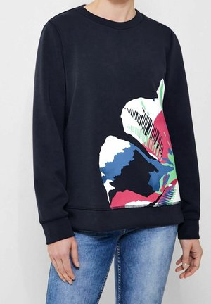 Persoon draagt een zwart shirt met lange mouwen met een grote meerkleurige abstracte bloemenprint aan de voorkant, gecombineerd met een blauwe spijkerbroek.